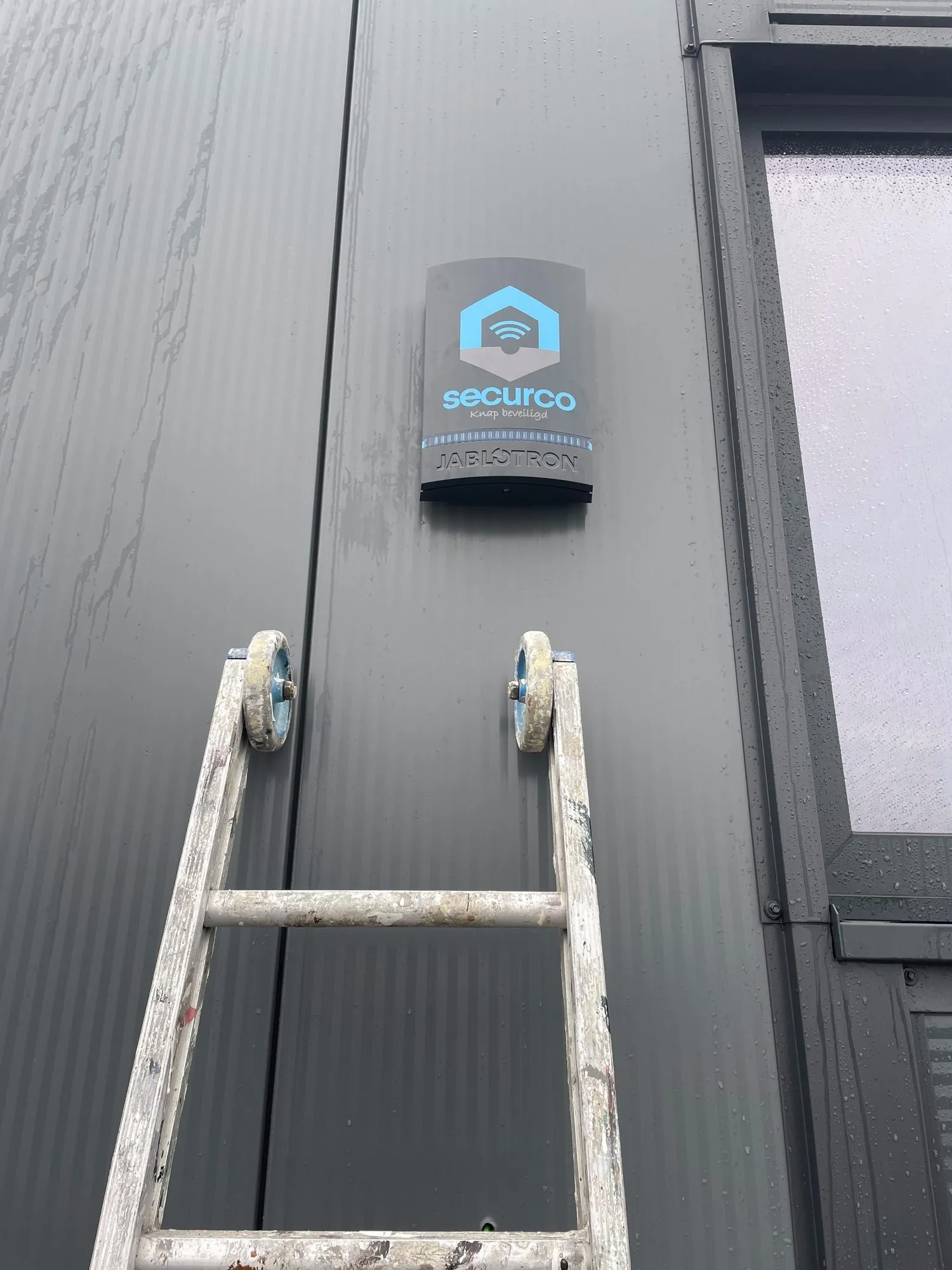 SecurCo beveiligingsbord met logo aan grijze wand, ladder ertegenaan