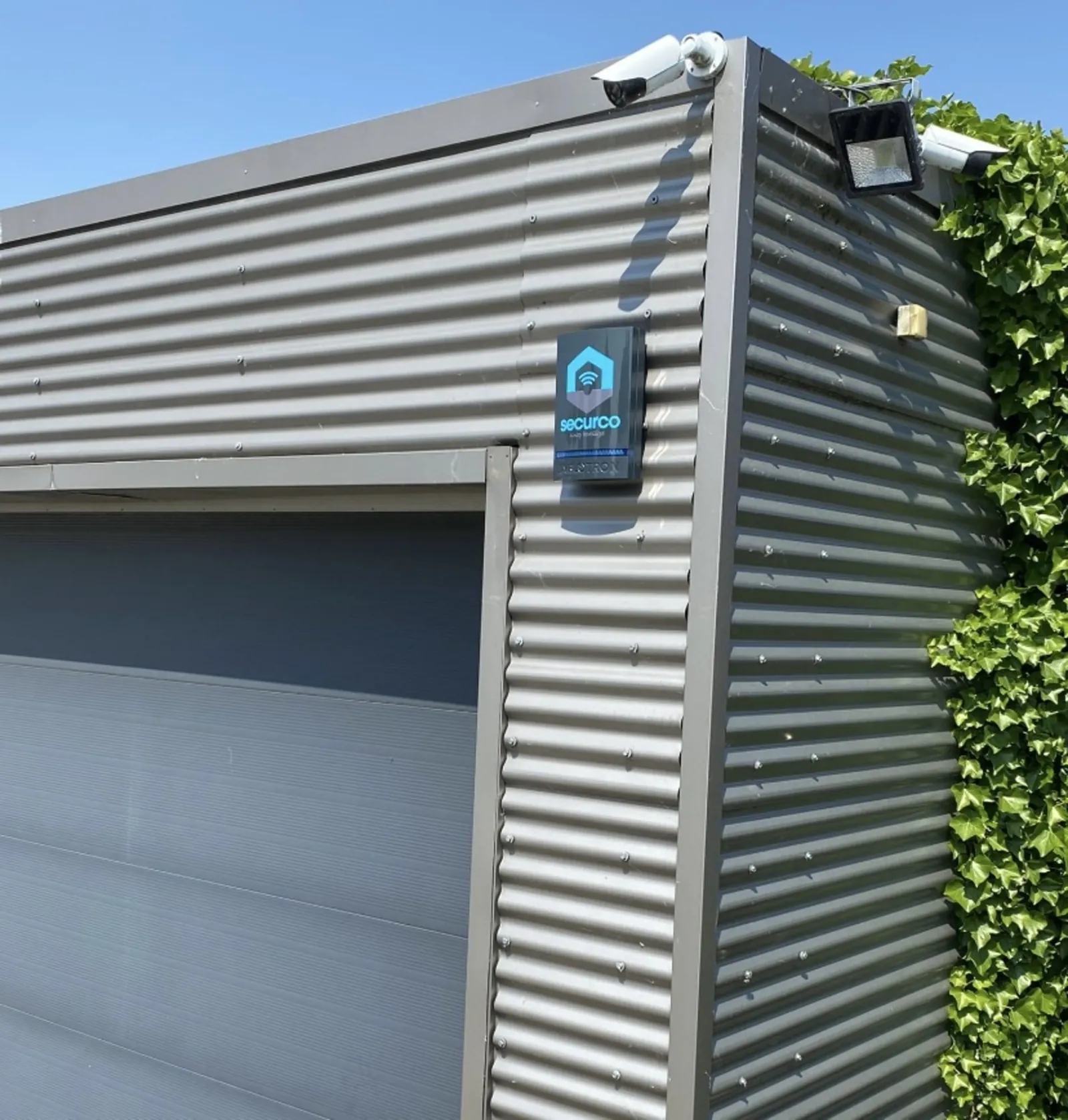 SecurCo beveiligingscamera en logo op grijze aluminium garagewand met groene haagplanten ernaast