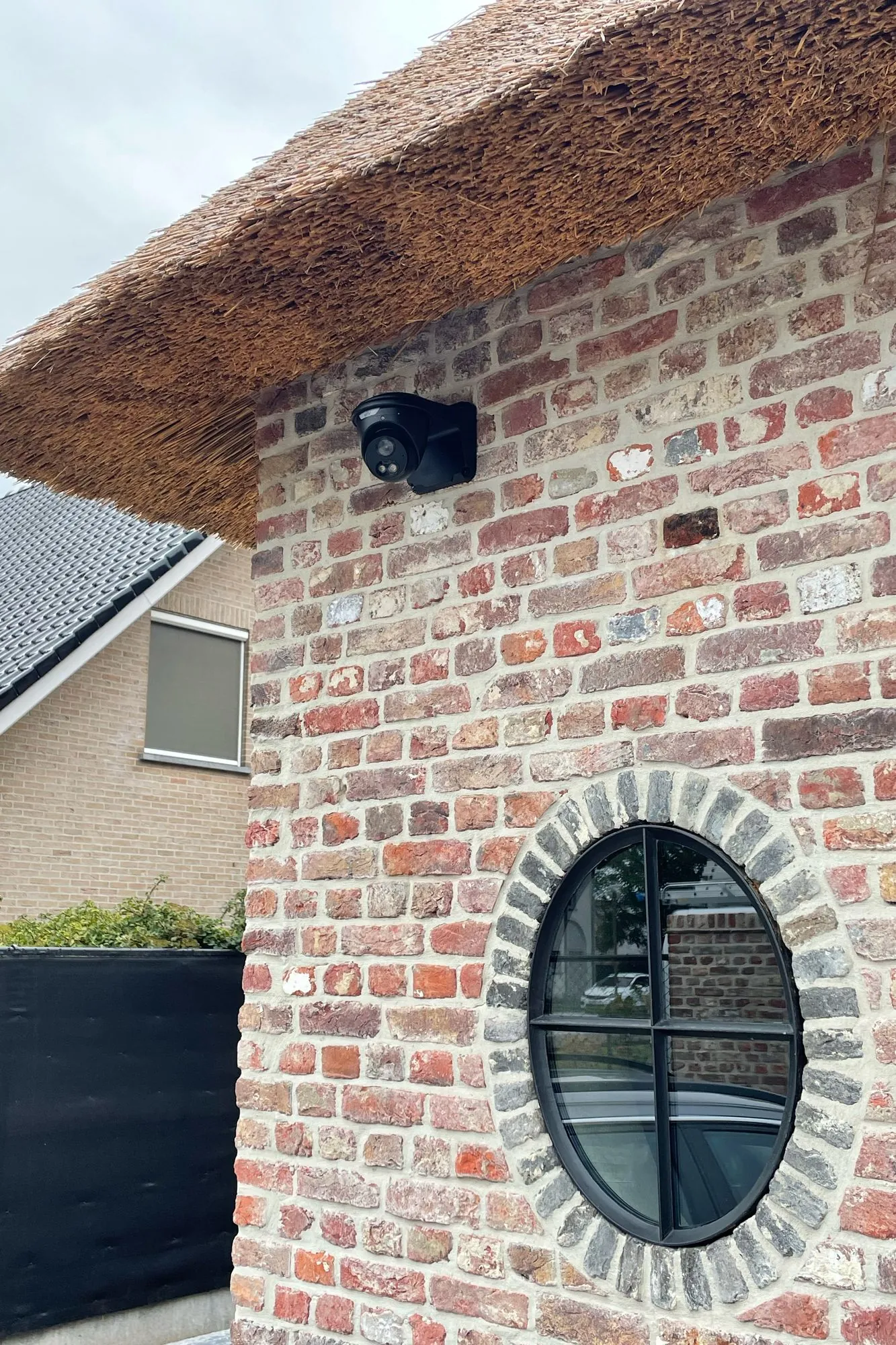 Zwarte beveiligingscamera gemonteerd op een bakstenen muur boven een ovaal raam van een landhuis