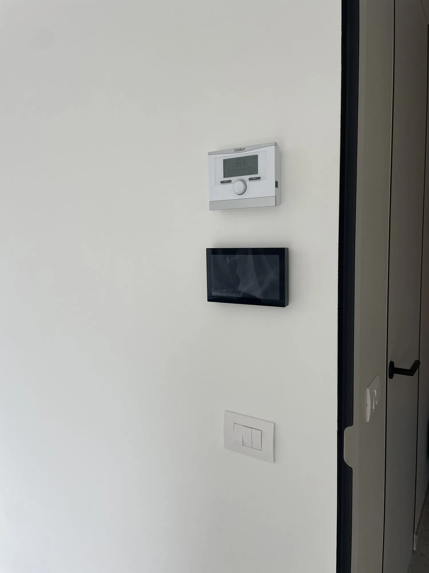 Witte thermostaat met display, zwart wandscherm en lichtschakelaar op witte muur naast donkere deur
